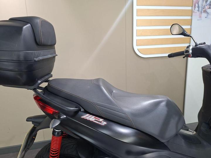 A close-up view of the rear section of a motorcycle, featuring a black seat with brown stitching and a matching rear storage compartment. The motorcycle has a visible red suspension spring and is set against a light-colored wall with decorative panels.