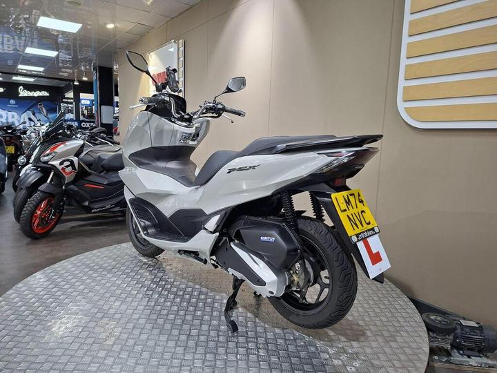 A new grey Honda PCX scooter displayed on a rotating stand in a motorcycle showroom, with various other scooters visible in the background.