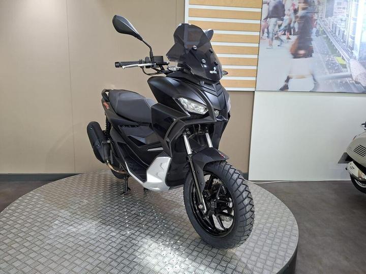 A sleek black scooter displayed on a circular metallic platform, featuring a windscreen, sporty design, and rugged tires, with a blurred cityscape in the background.