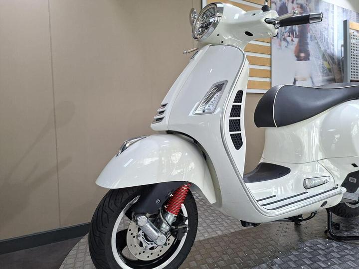 A white scooter positioned at an angle, showcasing its front wheel, chrome details, and black seat with white piping, set against a neutral background with soft lighting.