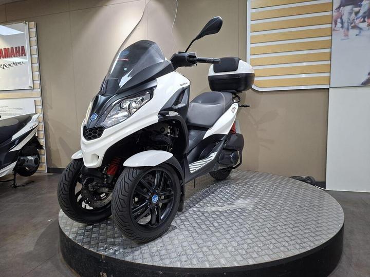 A white and black three-wheeled scooter displayed on a circular platform inside a showroom, featuring a windshield, sporty wheels, and a storage compartment at the rear.