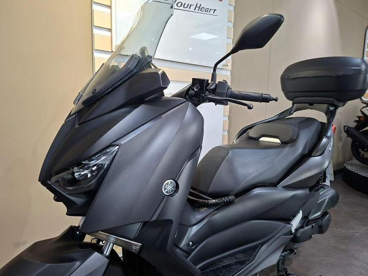 A matte black Yamaha scooter with a large windshield and comfortable seating, displayed in a showroom with light-colored walls.