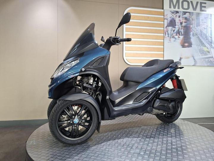 A blue three-wheeled scooter displayed on a round platform against a plain background with a sign that says "MOVE."