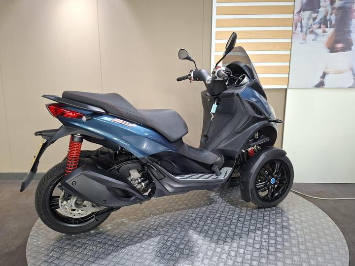 A three-wheeled scooter displayed on a circular, textured platform, featuring a sleek blue body, sporty red suspension, and a modern design with a windshield and dual headlights. The background showcases a simplistic interior with a light-colored wall and a blurred lifestyle image.