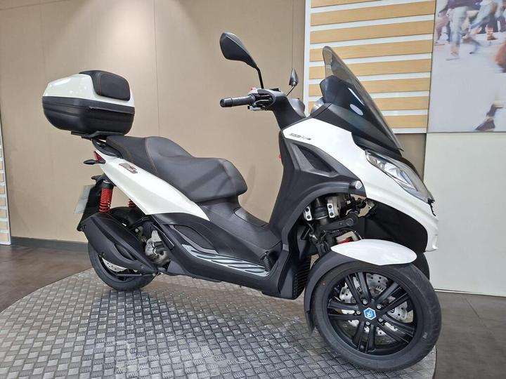 A three-wheeled scooter in white and black, featuring a large rear storage box, positioned on a textured surface with a blurred background.
