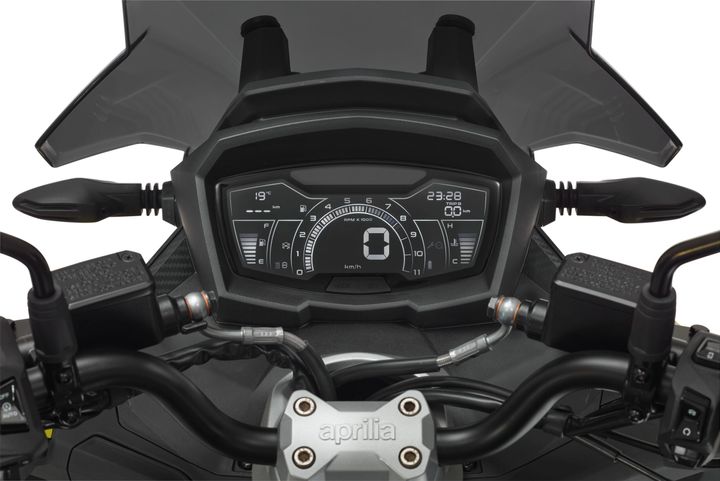 Close-up view of a motorcycle dashboard displaying gauges and indicators, with the Aprilia logo on the handlebar clamp, flanked by the handlebars and rearview mirrors.