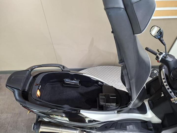 A top-down view of a scooter's seat with the seat raised, revealing a storage compartment beneath, along with a small black box inside the compartment and controls on the right. The background features a smooth surface and a plain wall.