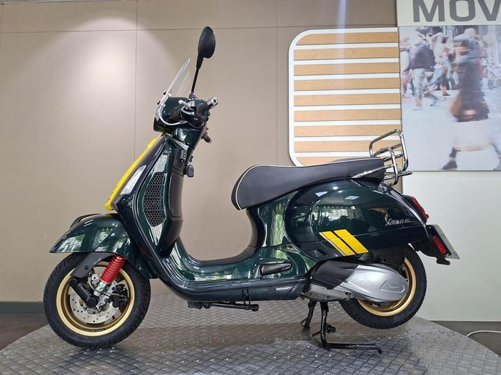 A green Vespa scooter with yellow accents, featuring a shield, rear rack, and red suspension, displayed on a patterned floor against a neutral background.