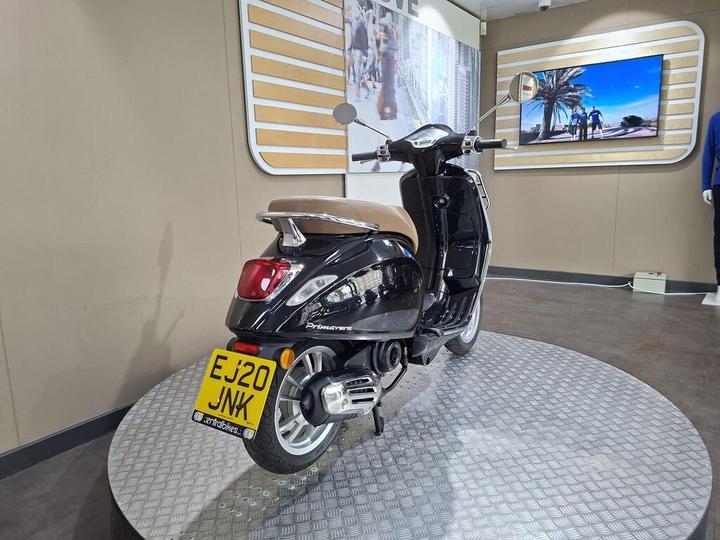 A black Vespa scooter with a brown seat, positioned on a round metal display platform in a showroom. A wall-mounted screen in the background displays an image of people walking.