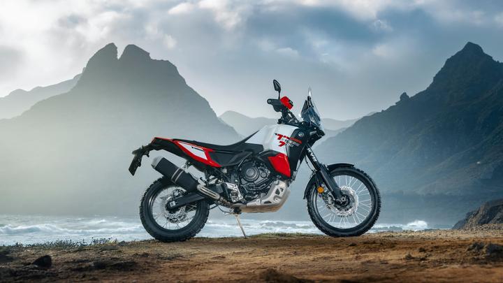 A motorcycle is parked on a rocky coastline with mountains in the background, under a partly cloudy sky.