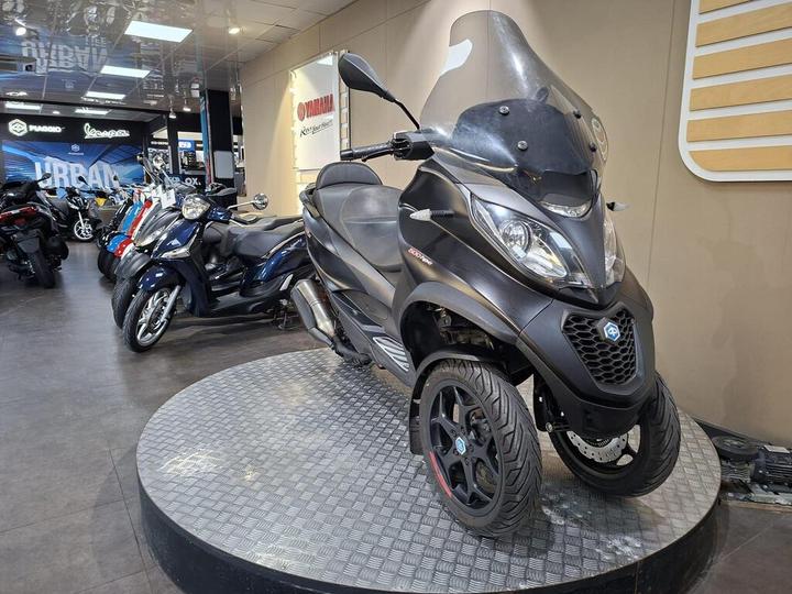 A black three-wheeled scooter displayed on a circular platform in a motorcycle showroom, with additional motorcycles visible in the background.