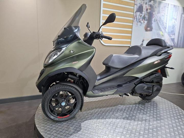 A green three-wheeled scooter with a windshield and black wheels on a round display platform, set against a light-colored backdrop.