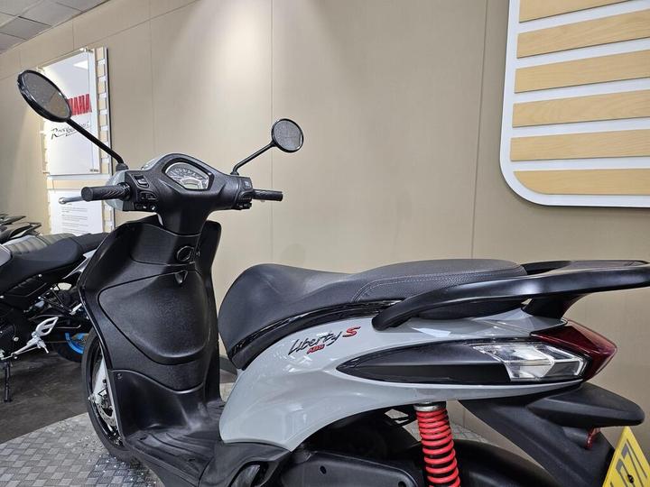 A close-up side view of a grey Yamaha Liberty S scooter, featuring a sleek design, black accents, and red suspension springs, displayed in a showroom setting.