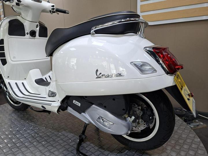 A sleek white Vespa scooter showcasing its side profile, with a smooth body design, black seat, and chrome accents, positioned on a diamond-patterned floor.