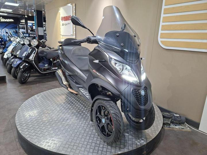 A black three-wheeled scooter displayed on a circular platform in a showroom, surrounded by other scooters in the background. The scooter features a large windshield and modern design elements.