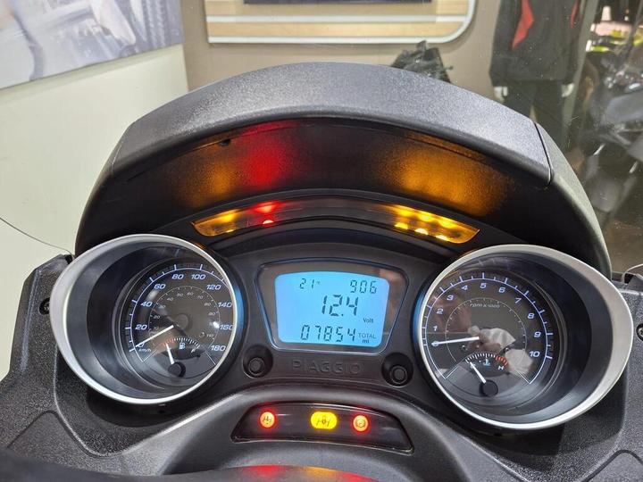 Dashboard of a Piaggio scooter displaying a speedometer on the left, a tachometer on the right, and a digital screen in the center with voltage and mileage readings. The background shows blurred images of other scooters.