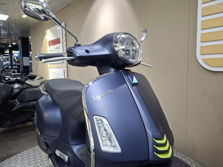 A sleek blue Vespa scooter is showcased in a modern display setting, featuring chrome mirrors, a circular headlight, and distinctive design elements such as an emblem and green accent near the front.