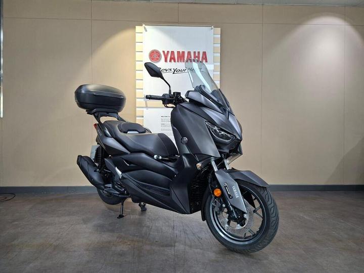 A sleek, dark-colored Yamaha scooter displayed in a showroom setting, featuring a prominent windscreen, large front wheel, and rear luggage compartment, with a Yamaha logo on the side and a branded sign in the background.