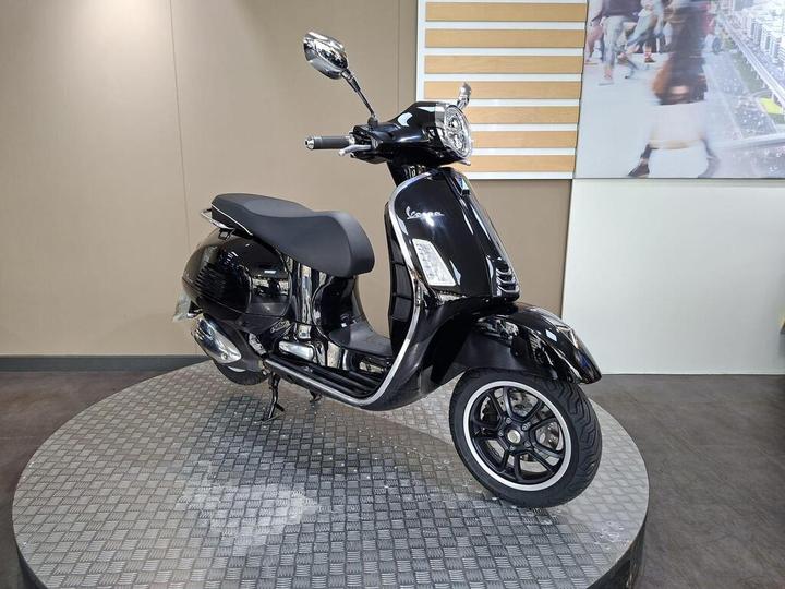 A sleek black scooter displayed on a round metallic platform, featuring a comfortable seat, chrome details, and modern design elements, against a minimalistic background.