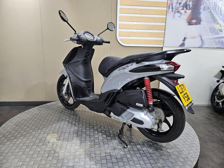 A silver and black scooter, model Vespa, is positioned on a textured circular platform with a simple background. The scooter is viewed from the rear-left angle, showcasing its sleek design and red shock absorbers.