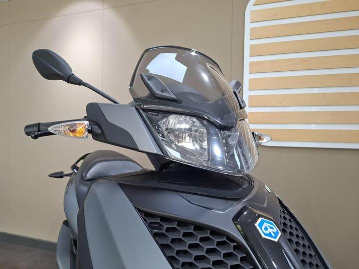 Close-up view of a scooter's front with a clear windscreen, headlight, turn signals, and handlebar reflected on a neutral background.