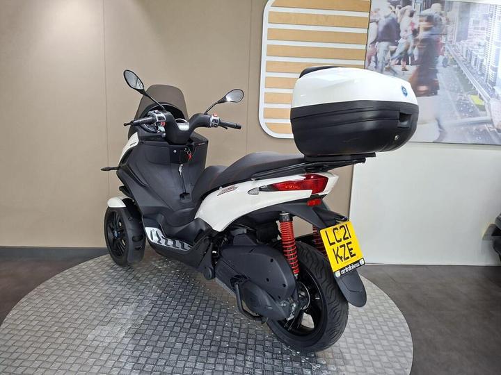A three-wheeled scooter in a showroom setting, featuring a black and white design with a rear storage box and a license plate visible. The scooter is positioned on a circular metallic platform against a neutral background.