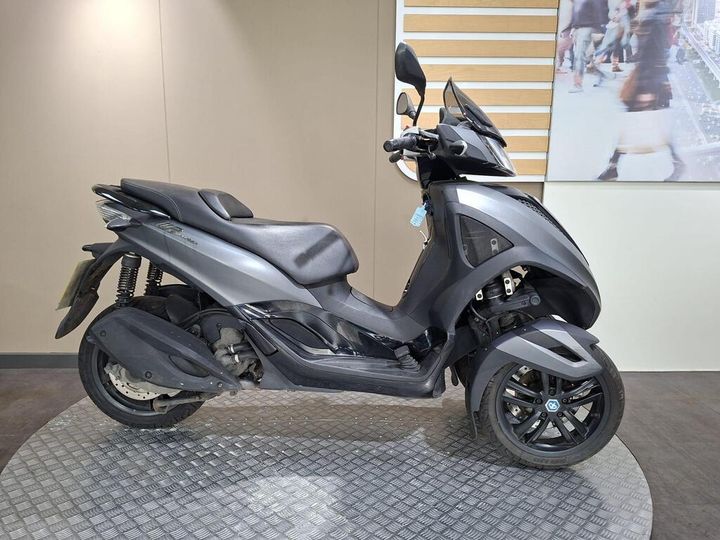 Piaggio MP3 300 Yourban LT (2018) A black and gray three-wheeled scooter parked on a textured circular platform against a light-colored wall.