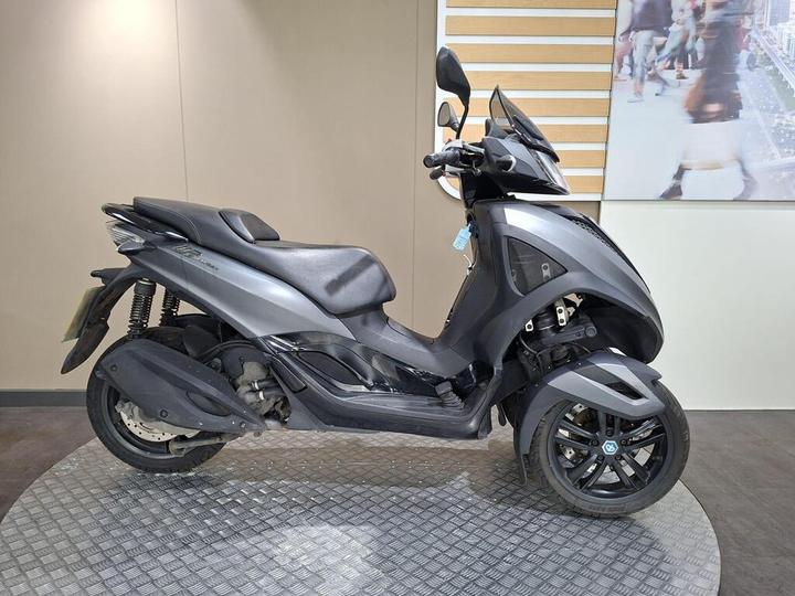 Piaggio MP3 300 Yourban LT (2018) A black and gray three-wheeled scooter parked on a textured circular platform against a light-colored wall.