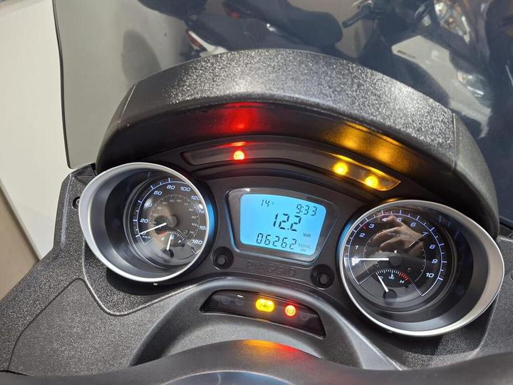 Close-up view of a scooter's dashboard displaying speedometer, tachometer, and digital readout with information on temperature, voltage, and mileage. Warning lights are illuminated.