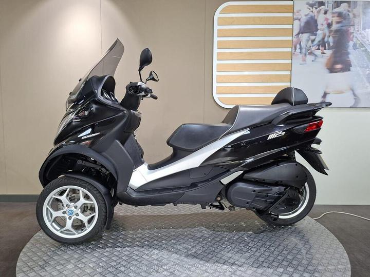 A black three-wheeled scooter displayed on a circular platform with a textured surface, featuring a large windscreen and a sleek design, set against a neutral wall background.