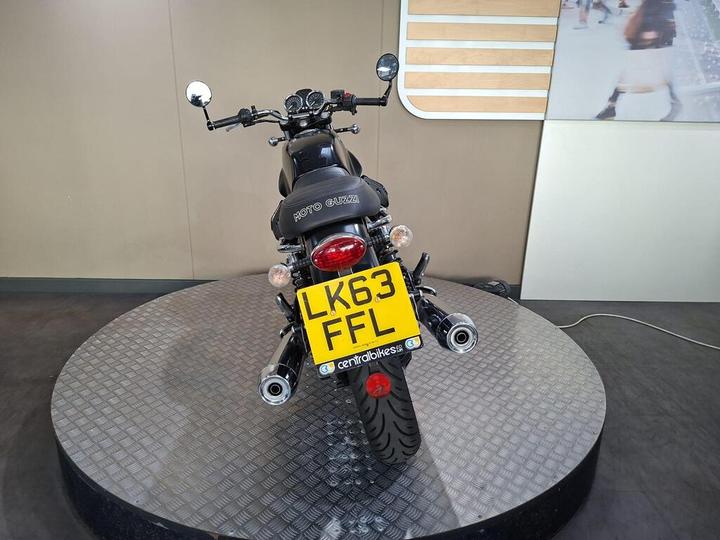 A black Moto Guzzi motorcycle displayed on a rotating circular platform, viewed from the rear. The motorcycle features a visible license plate (LK63 FFL) and has a sleek design with chrome elements. The background includes neutral-colored walls and a partial image on the right.