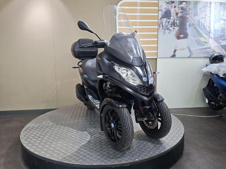 A black three-wheeled scooter displayed on a circular platform in a showroom, featuring a windshield and a storage compartment on the rear. A blurred image of people walking in the background.