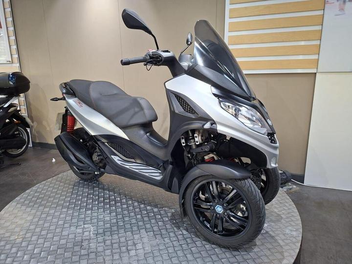 A silver three-wheeled scooter with a streamlined design and a tinted windshield, displayed on a textured platform, with another scooter visible in the background.