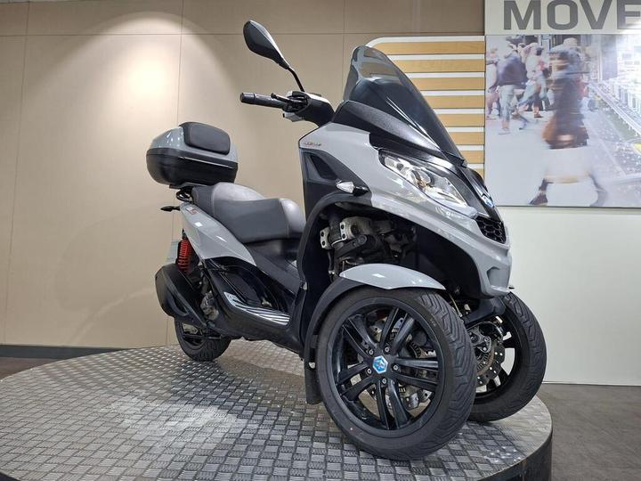 A gray three-wheeled scooter displayed on a round platform, featuring a large windshield, a rear storage compartment, and stylish black wheels. In the background, there's an abstract image of people walking.