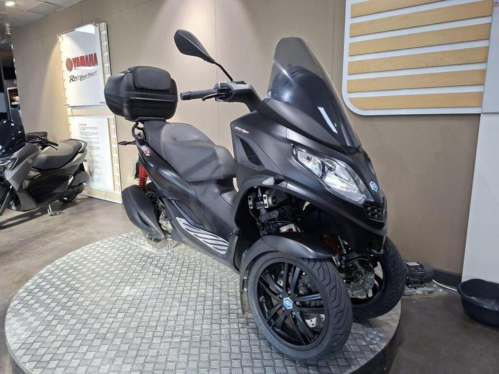 A sleek black three-wheeled scooter displayed on a circular metal platform, with a prominent windshield, comfortable seat, and a storage compartment on the back, set against a light-colored wall with branding.