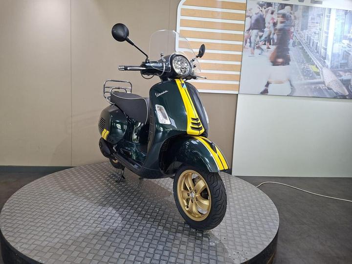 A vintage-style Vespa scooter in dark green with yellow accents, displayed on a round metallic platform. The backdrop features a blurred image of a busy street scene.