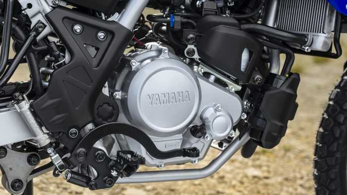 Close-up of a Yamaha motorcycle engine, showcasing the silver engine casing, black protective parts, and various components including pipes and bolts, set against a blurred natural background.