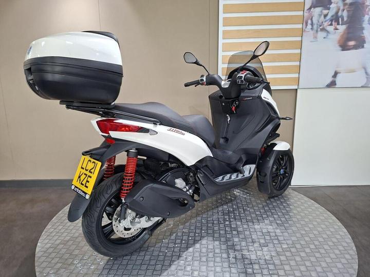 A white and black scooter with a rear storage compartment is displayed on a circular patterned platform, set against a neutral background with a blurred image of people. The scooter features red shock absorbers and a license plate visible at the rear.