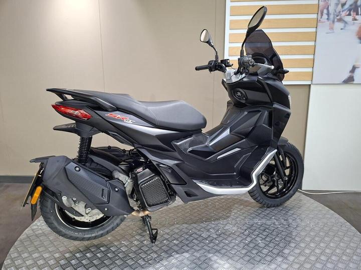 A black scooter with a modern design, featuring a prominent seat, rear tail light, and sleek bodywork, displayed on a circular metal platform against a neutral background.