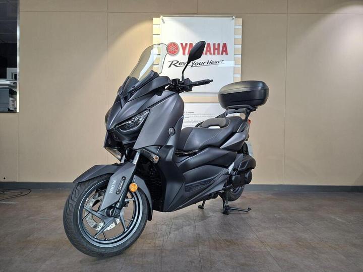 A sleek black Yamaha scooter is displayed in a showroom, featuring a prominent front headlight and a rear storage box, against a neutral background.