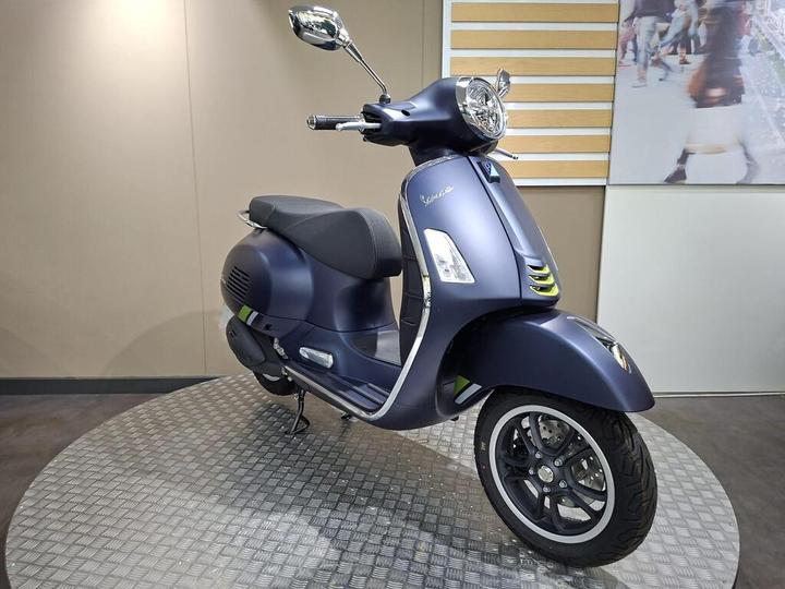 A blue matte Vespa scooter displayed on a circular metal platform against a beige wall backdrop. The scooter has a modern design with a distinctive front headlight and sleek lines.