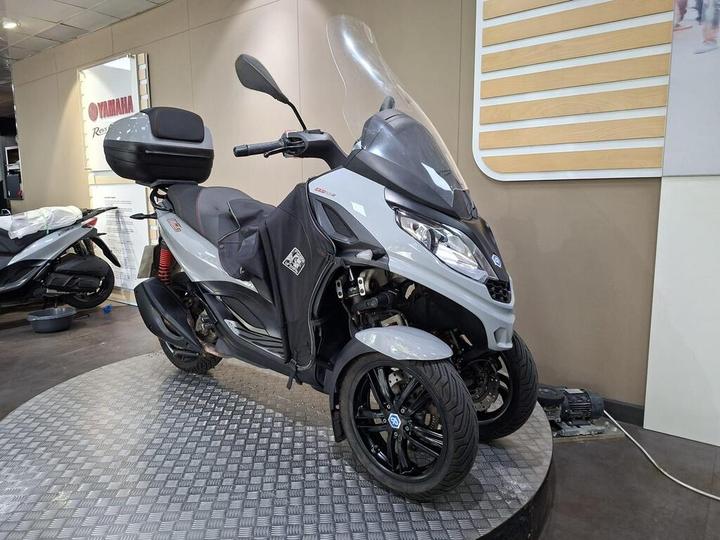 A gray and black three-wheeled scooter displayed in a showroom, featuring a large windscreen, a rear storage box, and a distinctive design. The background includes other scooters and a promotional wall for Yamaha.