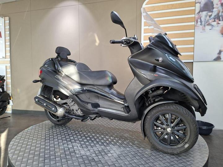 A black three-wheeled scooter is showcased on a round display platform in a well-lit indoor setting. The scooter features a sleek design, a tinted windshield, and a comfortable seat. In the background, there are neutral-colored walls and a blurred image of people.