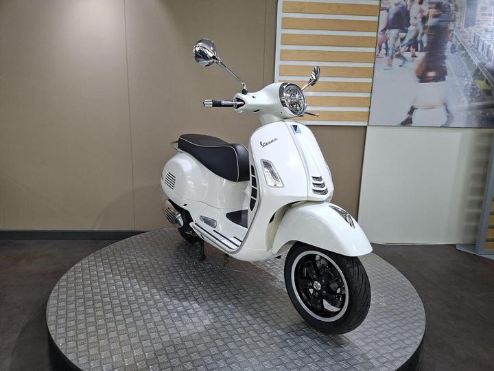 A white Vespa scooter displayed on a circular metal platform, featuring a black seat and chrome mirrors, with a blurred backdrop showing people and city scenery.