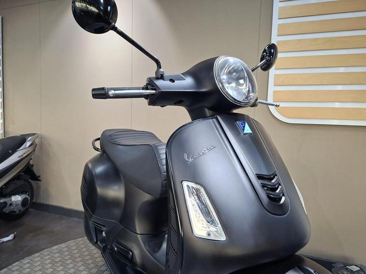 A black Vespa scooter with a sleek design is positioned in a modern indoor showroom, featuring a shiny headlight and rearview mirrors, with another scooter partially visible in the background.
