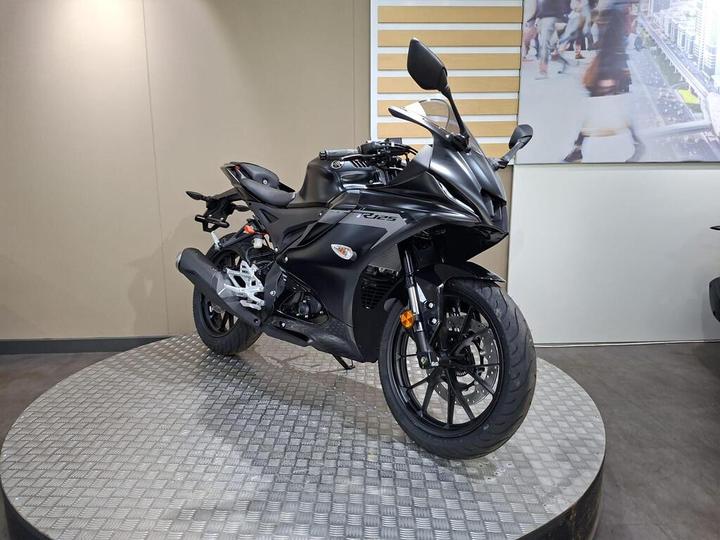A sleek black motorcycle displayed on a circular metallic platform, with a minimalistic backdrop featuring a blurred image of people walking.