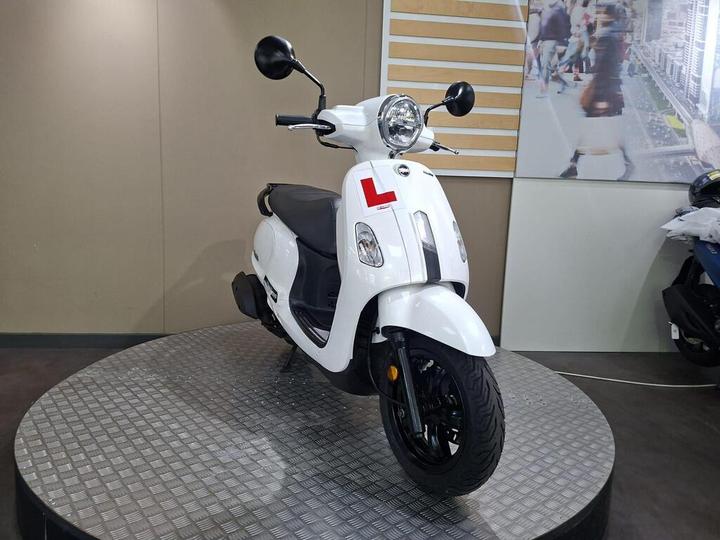 A white scooter on a rotating display stand, featuring a prominent "L" learner plate, set against a neutral background with a blurred urban scene.