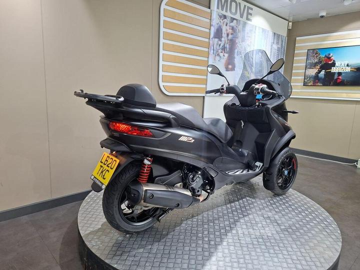 Side view of a modern black scooter displayed on a circular platform, featuring a sleek design, red shock absorbers, and a prominent windshield. The background includes a beige wall with decorative wooden panels and a screen showcasing motorcycle-related content.