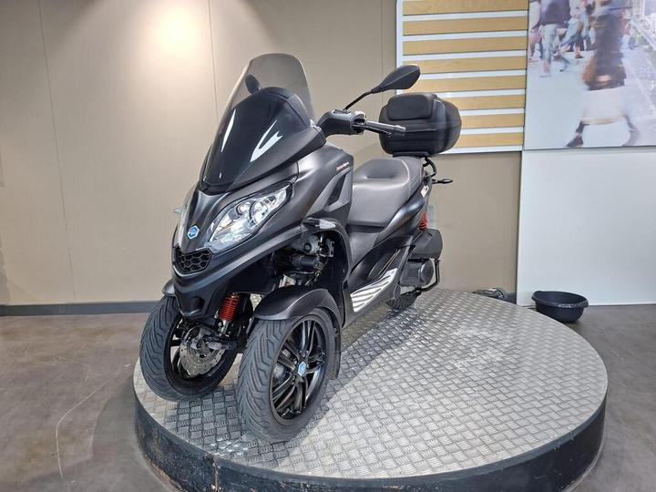 A black three-wheeled scooter is displayed on a textured circular platform, featuring a windshield, a comfortable seat, and visible storage at the rear. The setting has neutral-colored walls with a partially visible image in the background.