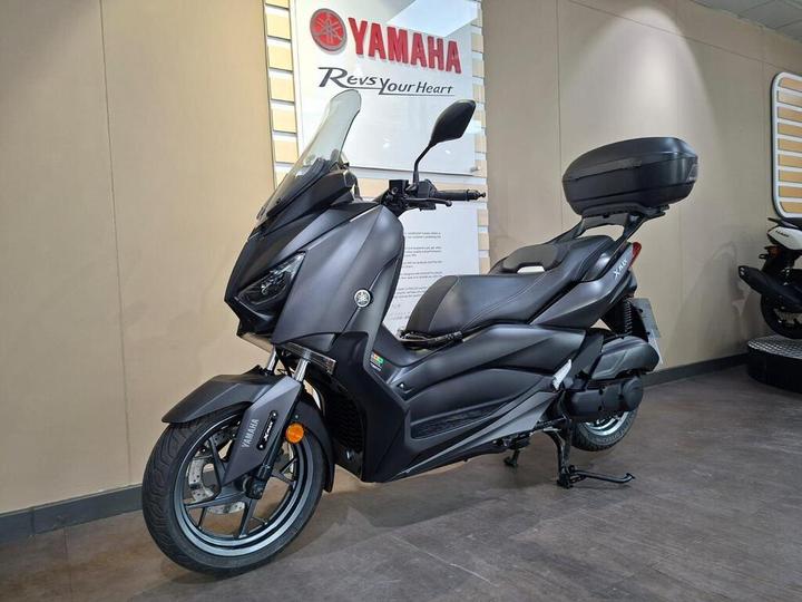A sleek black Yamaha scooter featuring a windshield, spacious seat, and rear storage box, positioned in a showroom with a Yamaha logo in the background.
